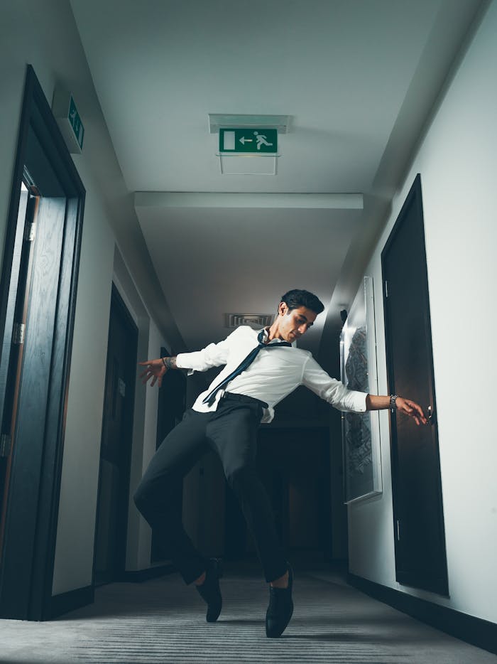 Stylish man striking a dance pose in a modern hallway with overhead lighting.