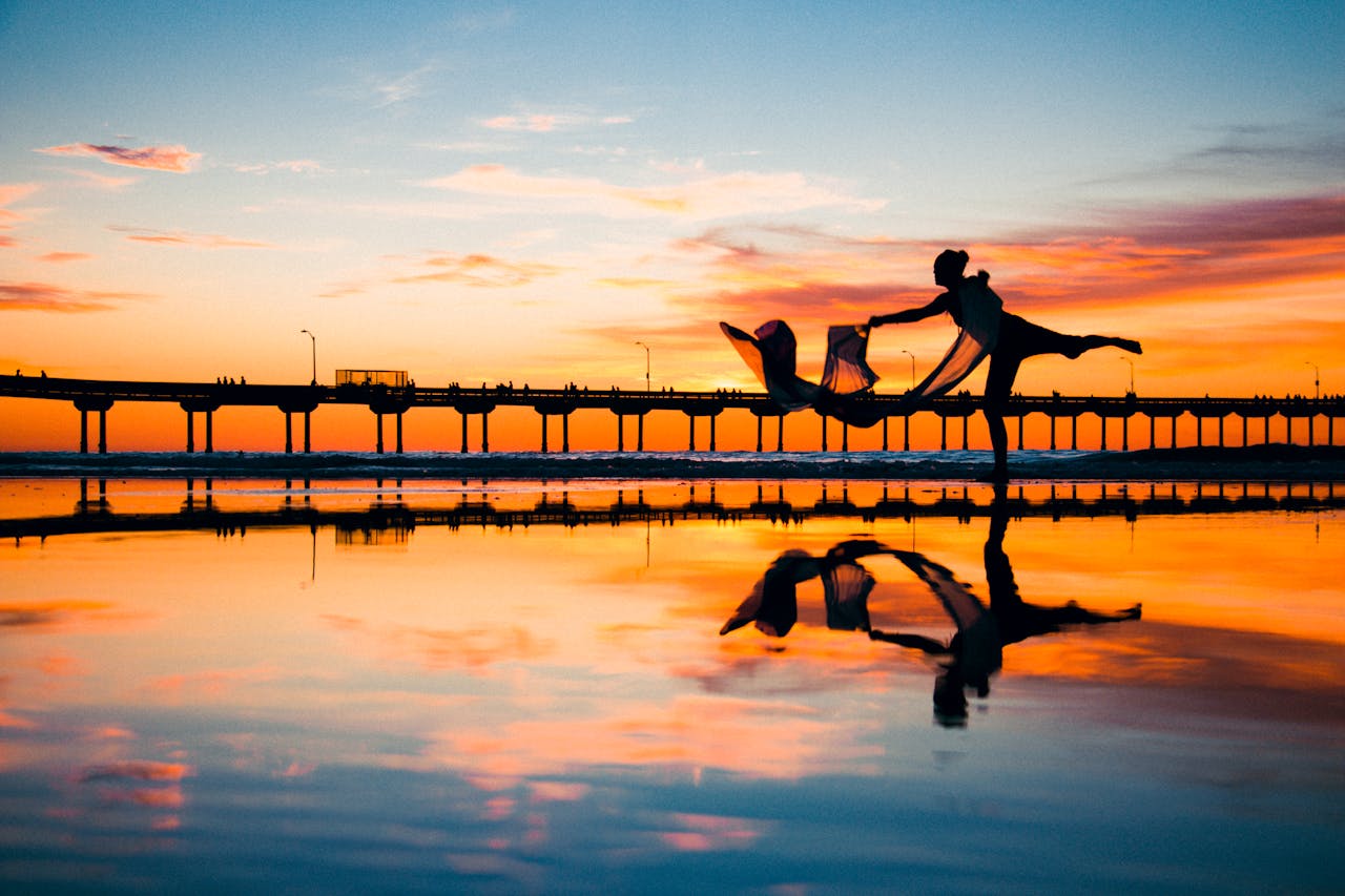 Silhouette of a dancer performing near a jetty at sunset with vibrant sky reflection.